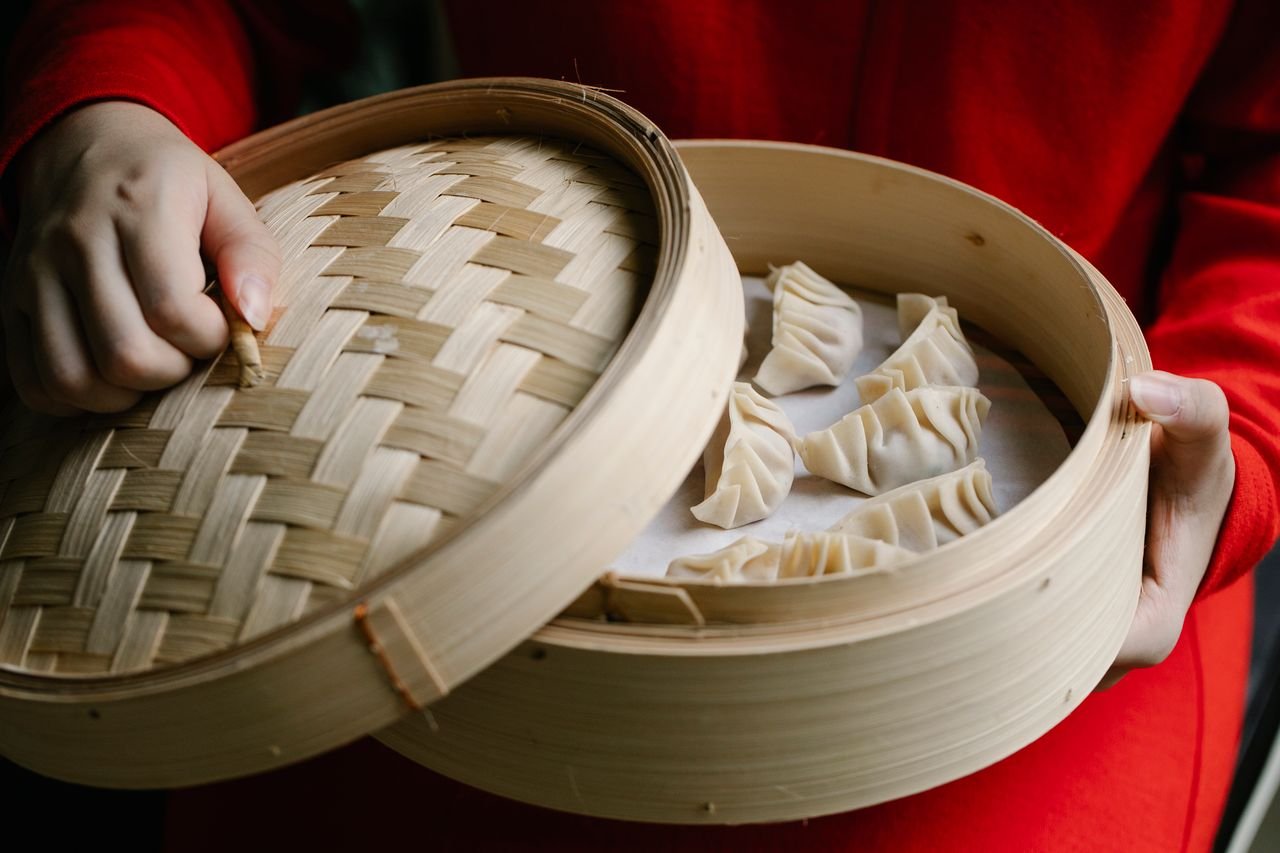 Crop anonymous cook in red clothes opening bamboo steamer with raw delicious dumplings